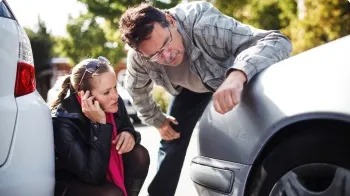 A couple check the damage to the front of their silver car while the woman calls her State Farm agent.